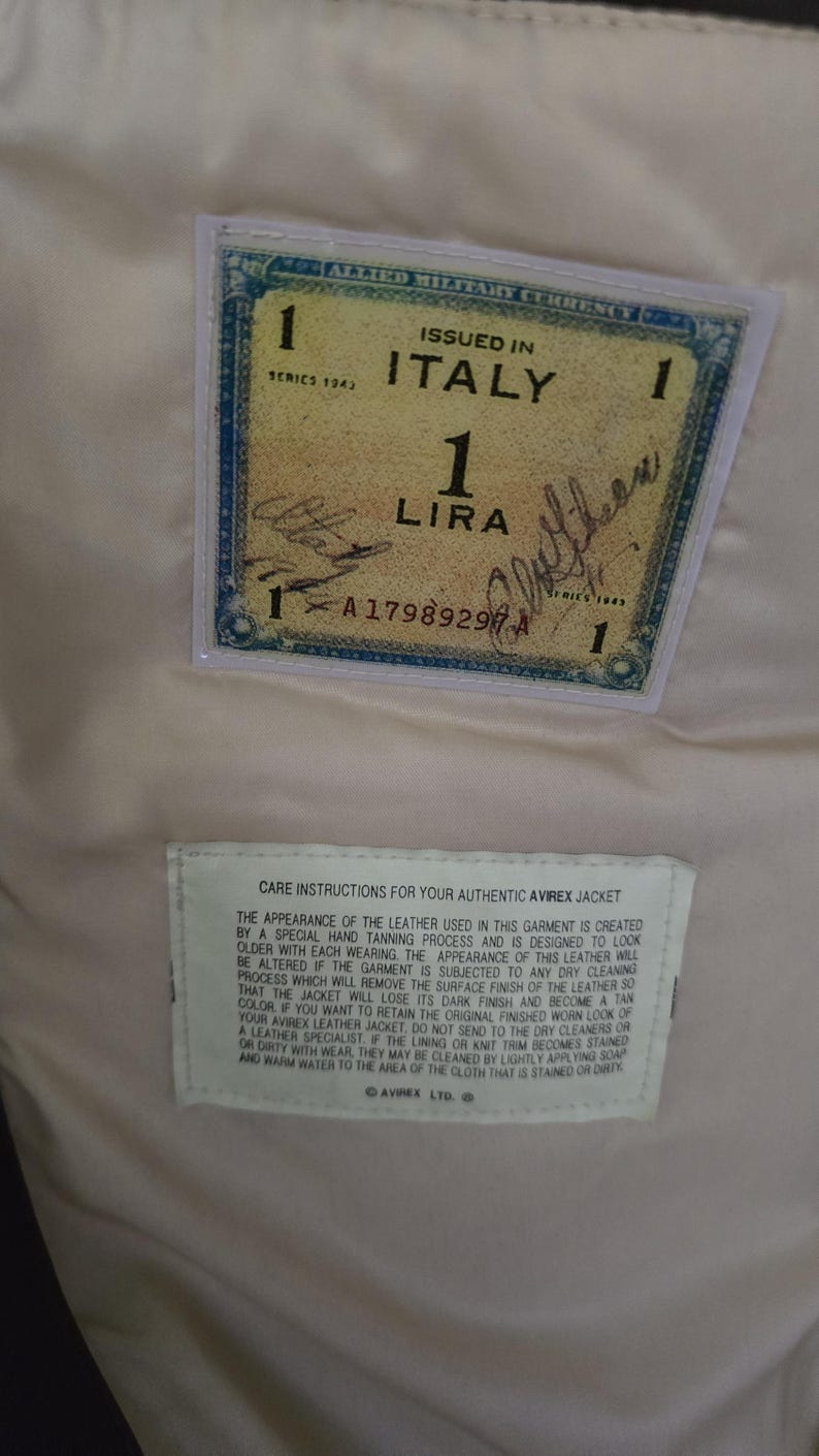 May include: Close-up of a vintage Allied Military Currency one lira note issued in Italy in 1943. The note is in a clear plastic sleeve, with a care instruction label below. The background is a light beige fabric.
