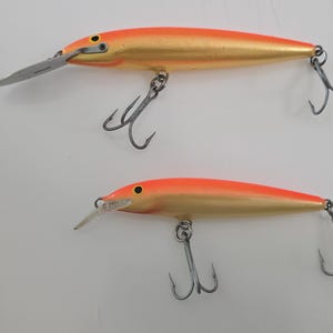 May include: Two orange and gold fishing lures with silver metal diving lips and two treble hooks each. The top lure measures approximately 5.5 inches in length, while the bottom lure measures approximately 4.5 inches in length on the ruler.