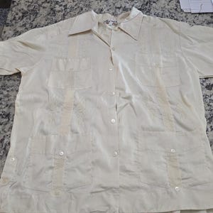 May include: Cream-colored short-sleeved shirt with a button-down front, collar, and four front pockets. The shirt has decorative embroidery down the front and on the pockets. The fabric appears to be a lightweight material.