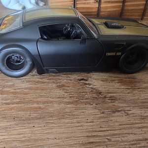 May include: Black and gold toy car with the text "TRANS AM" on the side. The car has a sleek design with large tires and a gold-colored roof. The car is sitting on a wooden surface.