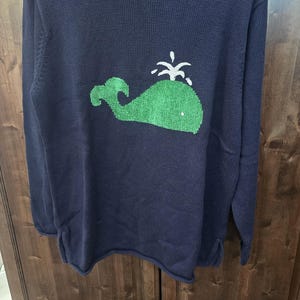 May include: A navy blue sweater with a green whale graphic. The whale is embroidered with white stitching and has a water spout.