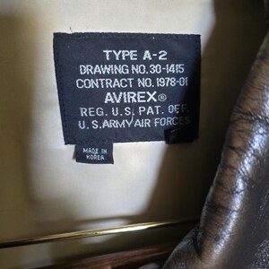 May include: Close-up of a black label inside a brown leather jacket. The label reads "TYPE A-2" and includes details such as "DRAWING NO.30-1415", "CONTRACT NO. 1978-01", and "AVIREX&reg;". The label also states "MADE IN KOREA".