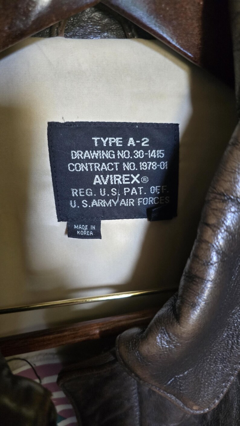 May include: Close-up of a black label inside a brown leather jacket. The label reads "TYPE A-2" and includes details such as "DRAWING NO.30-1415", "CONTRACT NO. 1978-01", and "AVIREX&reg;". The label also states "MADE IN KOREA".