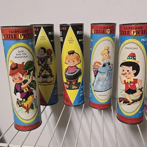 Può includere: Cinque contenitori cilindrici vintage Fairyland Puzzle Picture. Ognuno presenta un'illustrazione di cartoni animati diversa: Jack e il fagiolo magico, Jerry Jester, L'il Dutch Boy, Cenerentola e Pinocchio. I contenitori hanno accenti blu, rossi e gialli.