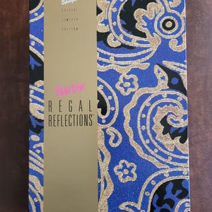 Puede incluir: Caja azul y dorada de Barbie "Regal Reflections", edición especial limitada. La caja presenta un panel vertical dorado con el logotipo y el texto de Barbie. El resto de la caja tiene un diseño de cachemira azul con detalles dorados.