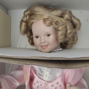 Puede incluir: Muñeca de porcelana con rostro sonriente y cabello rubio rizado, presentada en una caja de cartón. La muñeca lleva un vestido rosa con cuello de encaje blanco y detalles de estrellas. Un bloque de espuma beige es visible en la caja.