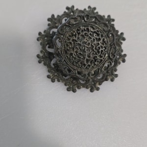 May include: Ornate, antique-style brooch with a dark, metallic finish. The brooch features a central circular design with intricate openwork, surrounded by a decorative, swirling frame. A vintage-inspired accessory.