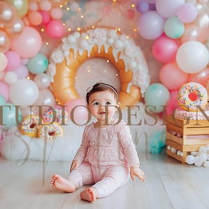 Könnte beinhalten: Ein Baby in einem rosa Spitzenoutfit sitzt vor einem bunten Ballonhintergrund. Der Hintergrund umfasst eine große donut-förmige Dekoration und eine kleine Holzkiste mit einem Donut-Kuchen. Der Text "STUDIO DESIGNS" ist sichtbar.