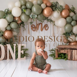May include: A baby sits in front of a backdrop with a balloon arch in shades of green, white, and copper. The word "one" is displayed in wooden letters. The image has the text "STUDIO DESIGNS".