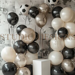 May include: A white cube sits in front of a wall decorated with black and white footballs and balloons. The balloons are black, white, and gold.