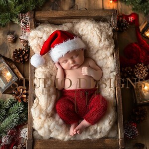 May include: A newborn baby wearing a red Santa hat and red knitted trousers is sleeping in a wooden crate filled with white fluffy material. The crate is surrounded by Christmas decorations, including pine branches, pinecones, and lanterns.