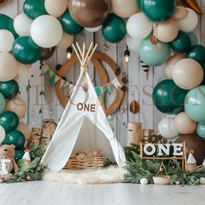 May include: A woodland-themed first birthday party setup with a white teepee tent, green and brown balloons, and a sign that says "ONE".
