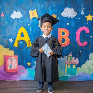 May include: A young child in a graduation gown and cap, holding a diploma, stands in front of a colorful backdrop. The backdrop features large letters "ABC", stars, clouds, and a painted scene. The child is smiling, celebrating a milestone.
