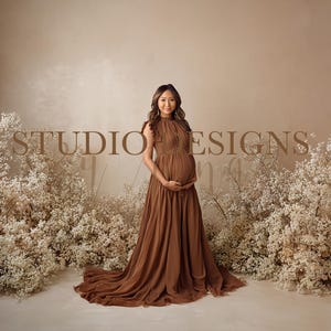 Puede incluir: Una mujer con un vestido de maternidad marrón fluido se encuentra frente a un telón de fondo con el texto "STUDIO DESIGNS". El vestido tiene un cuello alto y una larga cola. Flores blancas y follaje enmarcan la escena, creando una estética suave y elegante.