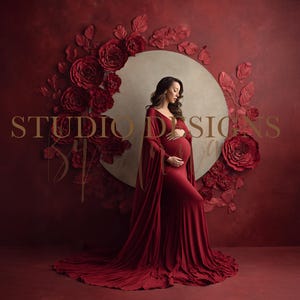 May include: A woman in a long, flowing red maternity gown stands before a burgundy backdrop. The backdrop features a large, circular floral arrangement with the words "STUDIO DESIGNS" in gold lettering.
