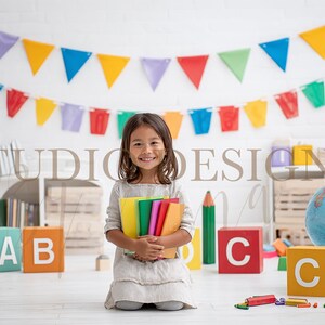 Könnte beinhalten: Ein junges Mädchen lächelt und hält bunte Bücher in einem Klassenzimmer. Die Szene umfasst Alphabetblöcke, einen Globus und ein Banner mit bunten Wimpeln. Ein großer Bleistift und der Text "STUDIO DESIGNS" sind ebenfalls sichtbar.