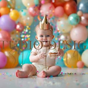 Könnte beinhalten: Ein Baby mit einer goldenen Krone sitzt vor einem bunten Ballonhintergrund und isst ein Cupcake. Konfetti liegt auf dem Boden verstreut.