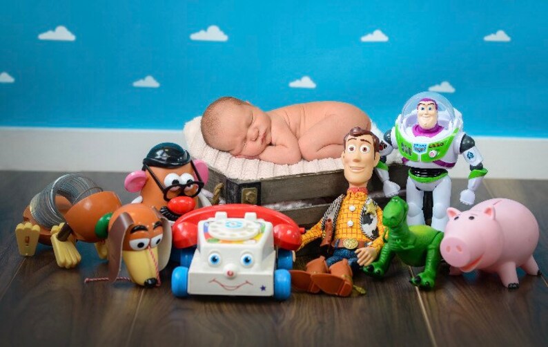 May include: A newborn baby is sleeping on a white blanket surrounded by a group of popular toy characters, including Woody, Buzz Lightyear, Mr. Potato Head, Slinky Dog, and a toy phone. The toys are arranged on a wooden surface.