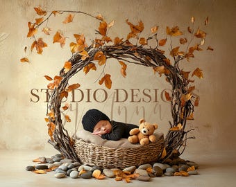 May include: A newborn baby in a black outfit and beanie sleeps peacefully in a woven basket, nestled inside a circular twig frame adorned with autumn leaves. A small teddy bear rests beside the baby. The scene is set on a neutral background.
