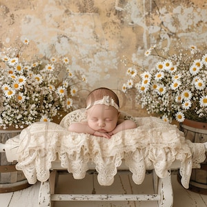 Puede incluir: Un bebé durmiendo con una diadema blanca yace en una cama de crochet blanco con un borde de encaje blanco. La cama está sobre una superficie blanca con dos barriles de madera llenos de flores blancas a cada lado.