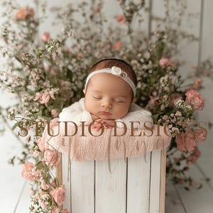 May include: A newborn baby wearing a white knitted sweater and a white headband with a pearl accent, sleeping in a pink blanket inside a white wooden box. The box is surrounded by pink flowers.