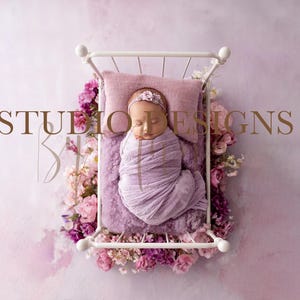 May include: A newborn baby wrapped in a lavender swaddle, lying on a lavender pillow and blanket inside a white metal bed frame. The bed is surrounded by pink and purple flowers. The image has the text "STUDIO DESIGNS" in gold.