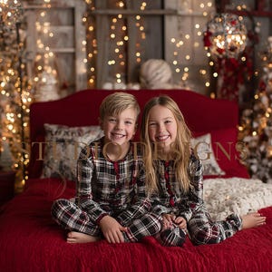May include: Two children in matching black, red, and white plaid pajamas sit on a red bed in front of a Christmas tree. The room is decorated with Christmas lights and ornaments, creating a festive holiday scene.
