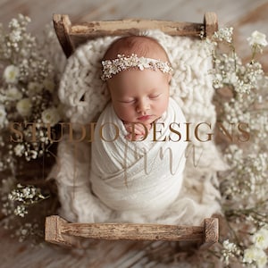 Puede incluir: Un bebé recién nacido envuelto en una manta blanca, con una diadema floral, yace en una pequeña cama de madera. La cama está sobre una superficie de madera y rodeada de flores blancas. El texto "STUDIO DESIGNS" está en el centro.