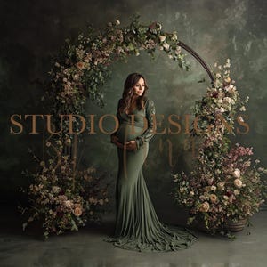 Pode incluir: Uma mulher em um vestido de maternidade verde-oliva está em frente a um arco floral. O arco é adornado com flores e vegetação, em um fundo verde escuro. As palavras "STUDIO DESIGNS" são visíveis.