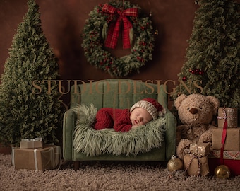 May include: A newborn baby wearing a red outfit and a Santa hat, sleeping on a green couch with a faux fur throw. The scene is decorated with Christmas trees, a wreath, a teddy bear, and wrapped gifts.