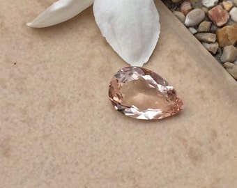 Puede incluir: Una piedra preciosa de color rosa claro en forma de pera con facetas. La piedra preciosa está sobre una superficie de color marrón claro.
