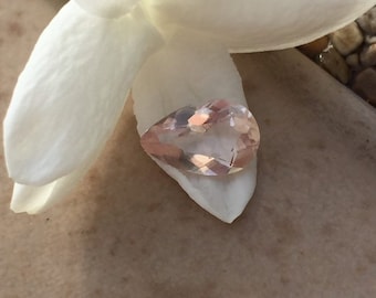 Puede incluir: Una piedra preciosa de color rosa pálido en forma de pera con facetas, descansando sobre un pétalo de flor blanco.