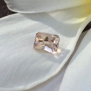 May include: A light pink, rectangular gemstone with a faceted cut. The gemstone is set against a white background.