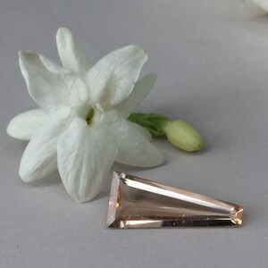 May include: A pale pink, faceted gemstone with a triangular shape. The gemstone is lying on a white surface next to a white flower.