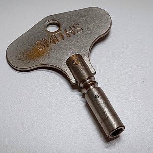 May include: A vintage metal clock key with the word "SMITHS" stamped on the head. The key has a cylindrical stem and a small hole near the head. The key is silver in color and shows signs of age.
