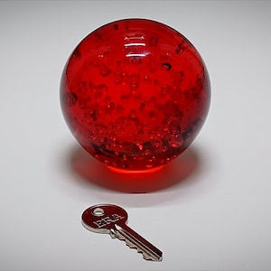 May include: A large, round, red glass orb with a textured interior, resting on a white surface. A silver key with the word "ERA" is positioned below the orb. The orb has a glowing effect.