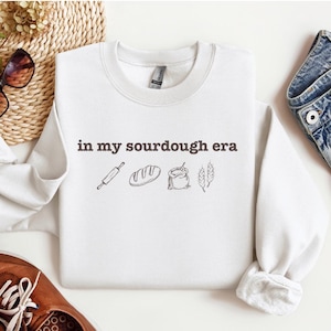 Può includere: Felpa bianca con la scritta "in my sourdough era" e illustrazioni di un matterello, una pagnotta di pane, un sacco di farina e spighe di grano.