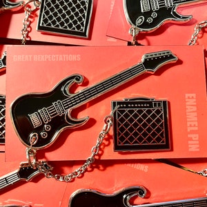 Può includere: Uno spillo smaltato a forma di chitarra elettrica nera e amplificatore con una catena argentata. Lo spillo è su uno sfondo rosso con il testo "Great Expectations" e "Enamel Pin".