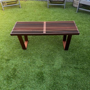 May include: A dark brown wooden bench with slatted top and a simple design. The bench is sitting on a green artificial grass lawn.