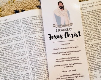 Puede incluir: Un marcapáginas blanco con una ilustración azul y blanca de Jesucristo. El marcapáginas tiene el texto "Because of Jesus Christ" y una lista de afirmaciones positivas sobre Jesucristo.