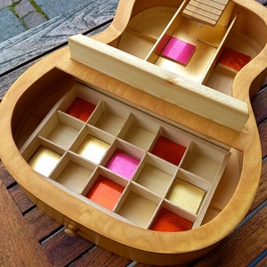 Könnte beinhalten: Eine gitarrenförmige Schmuckschatulle aus Holz mit mehreren Fächern. Der Innenraum verfügt über kleine, quadratische Fächer mit goldenen, rosa und roten Akzenten. Die Box ist geöffnet und zeigt die Stauräume, und steht auf einem Holztisch.
