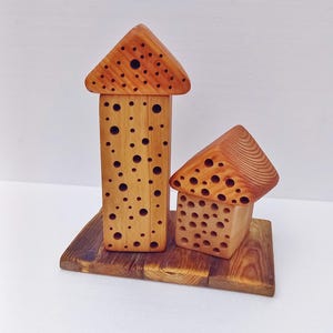 May include: Two wooden house sculptures with triangular roofs and numerous circular cutouts. The houses are light brown and sit on a rectangular wooden base. The taller house is vertical, while the smaller one is angled.