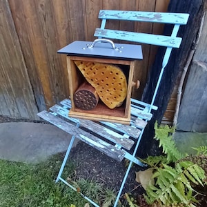 May include: A wooden insect house with a gray lid and a metal handle. The house is filled with different types of nesting materials for insects, including hollow logs and drilled holes. The house is sitting on a blue folding chair.
