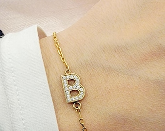 May include: Gold-toned bracelet featuring the letter "B" encrusted with small, clear gemstones. The bracelet has a delicate chain design and is worn on the wrist. The jewelry is set against a soft, neutral background.
