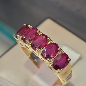 Puede incluir: Un anillo de oro con cinco rubíes de corte ovalado. Los rubíes son de color rojo intenso y están engastados en una banda de oro. El anillo se muestra sobre una superficie neutra.
