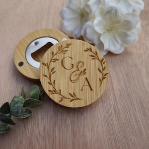 May include: A wooden bottle opener with a silver opener and a round wooden coaster. The coaster is engraved with the letters "G & A" inside a laurel wreath. White flowers and greenery are in the background.