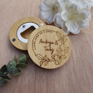 May include: A bamboo bottle opener with a silver metal opener and a round wooden magnet. The magnet features the names "Andreja & Tadej" and the date "25.5.2024" engraved within a floral wreath. The opener is made of bamboo.