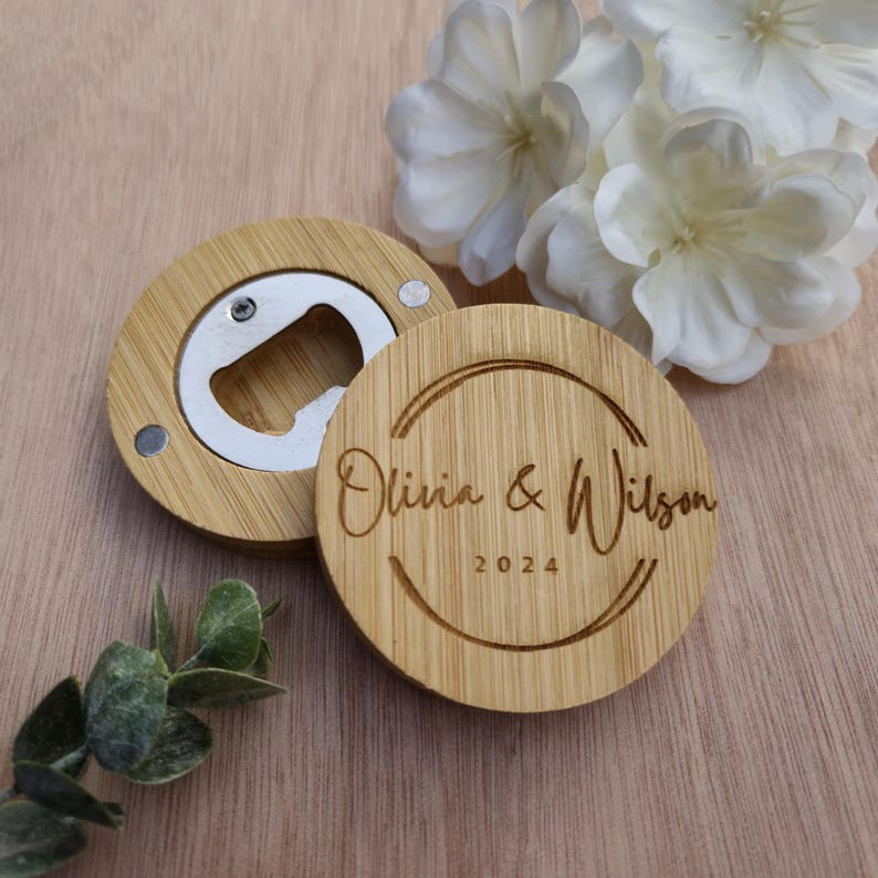 May include: Two round wooden bottle openers with a silver opener and magnetic closure. One opener has the names "Olivia & Wilson" and the year "2024" engraved. The openers are on a light brown surface with white flowers.