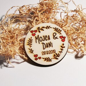 Può includere: Un cartello rotondo in legno con un design a corona di fiori. Il cartello è decorato con cuori rossi e il testo "MOJCA & DANI 28.9.2019".