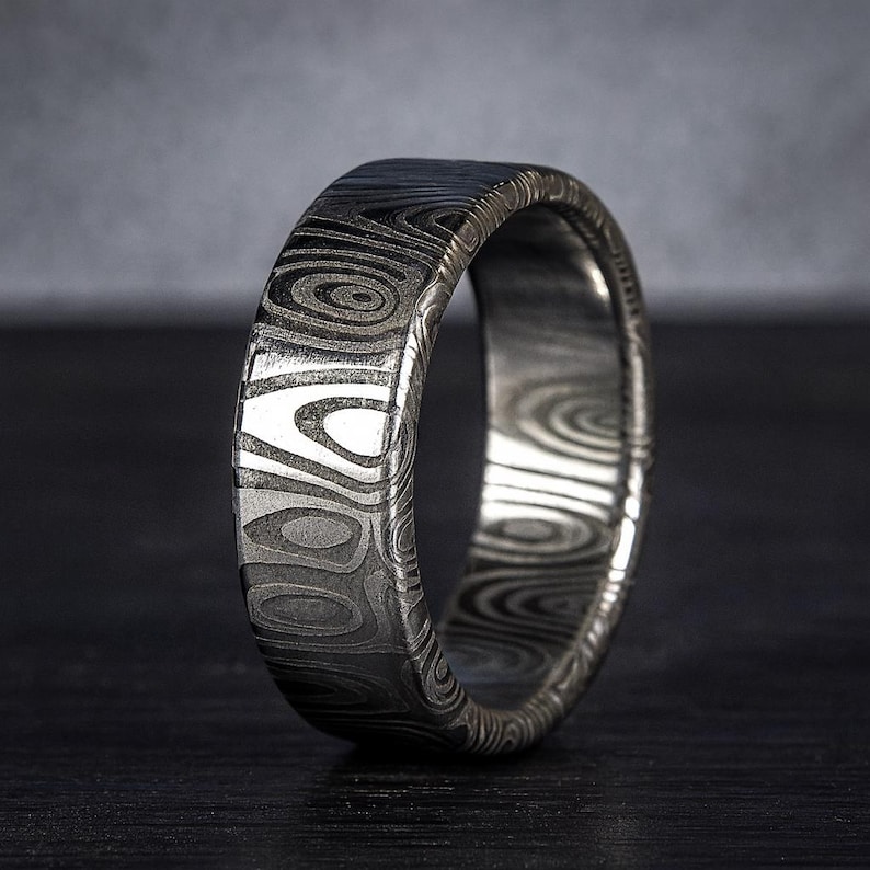 Puede incluir: Primer plano de un anillo plateado con un patr&oacute;n &uacute;nico. El anillo tiene una superficie texturizada con dise&ntilde;os org&aacute;nicos y arremolinados. El anillo es de metal y tiene un acabado pulido. Es una joya.